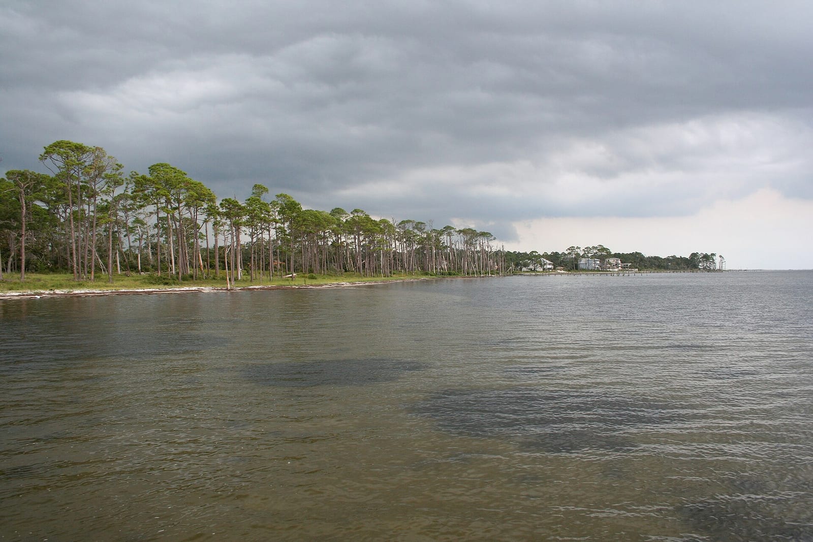 Santa Rosa Sound shoreline at Navarre with a low sandy bank lined with pines, waterfront homes and a dock in the distance, under a heavy gray storm sky