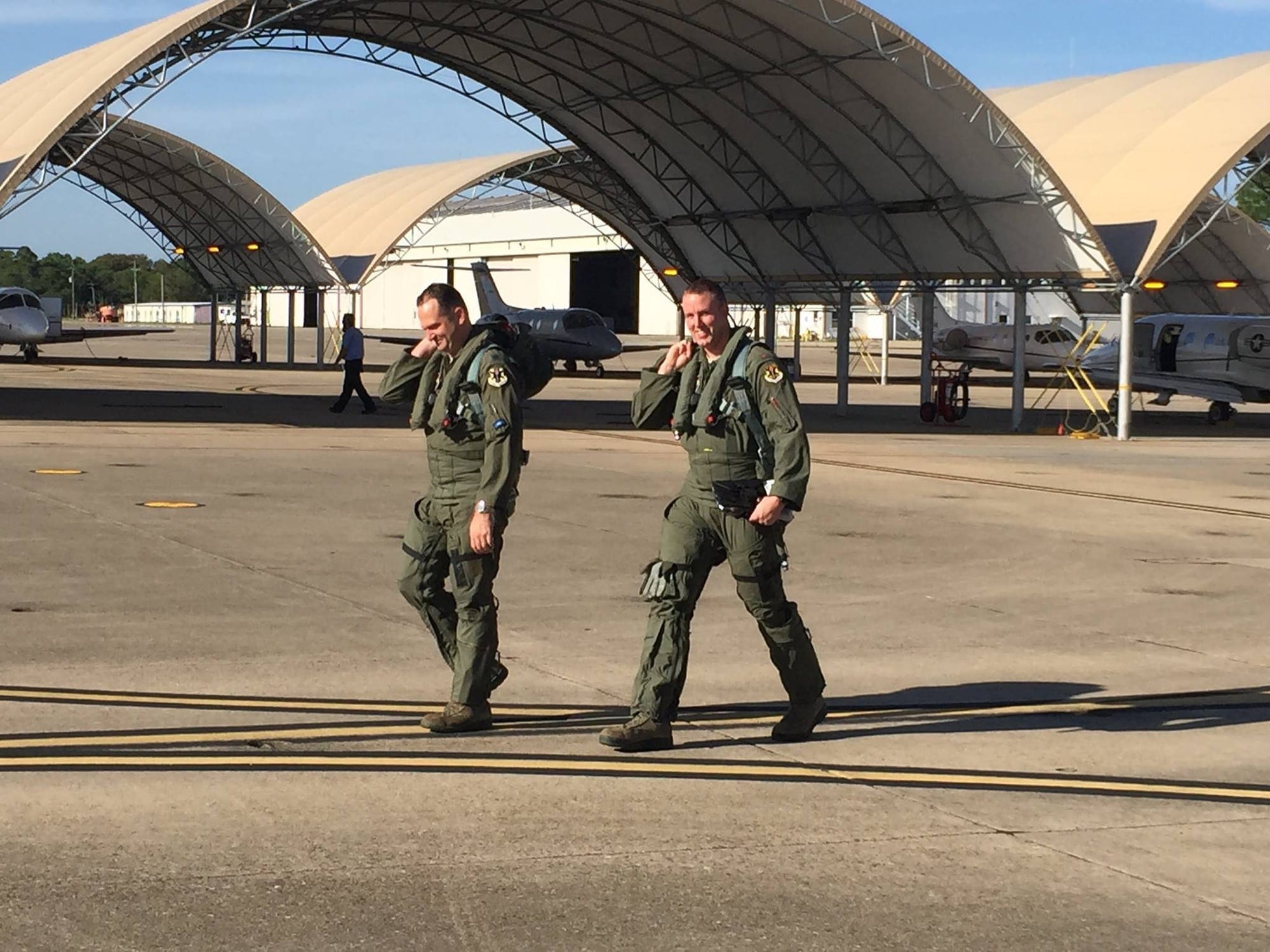 Two aviators in flight suits walking across a flightline in front of sunshade hangars and trainer aircraft